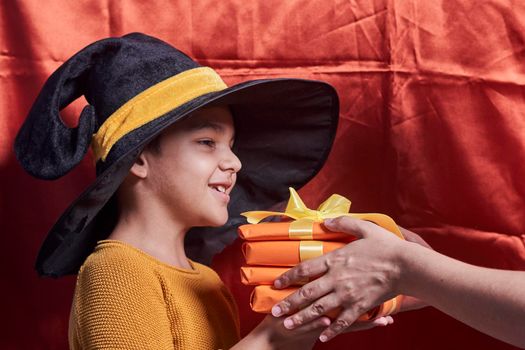A little kid in witch cap holding a pile of gift boxes. Halloween gifts in hands of a boy over orange background