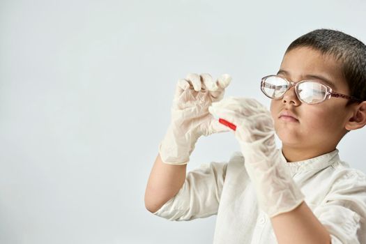 A portrait of a cute boy in protective gloves and glasses holding a piece of red plasticine in hands against the white background