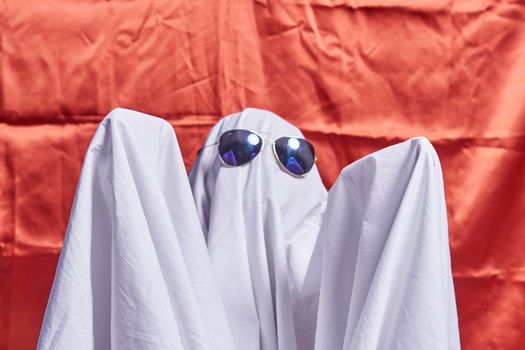White ghost on orange. Halloween holiday. A young child dressed in a ghost costume for Halloween on orange background. Funny Halloween kid. Little cute child in white costume and sunglasses