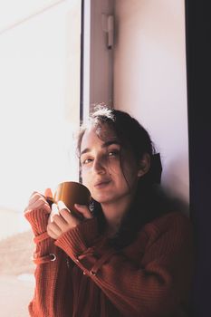 Cold autumn days - a young multi-racial female drinks coffee in a cozy windowsill. Middle-eastern or mixed-race woman enjoys weekend drinking hot tea near the windows