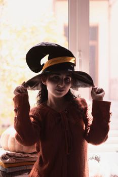 Halloween lady wearing witch cup. A woman in witch hat and Halloween pumpkin near the windows. Portrait of young female in orange sweater and traditional black witch cap