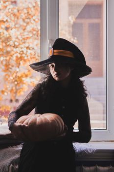 Halloween lady wearing witch cup. A woman in witch hat and Halloween pumpkin near the windows. Portrait of young female in orange sweater and traditional black witch cap