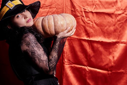 Halloween lady wearing witch cup and holding a big Halloween pumpkin. A woman in witch hat and Halloween pumpkin over orange background. Portrait of young female in traditional black witch cap