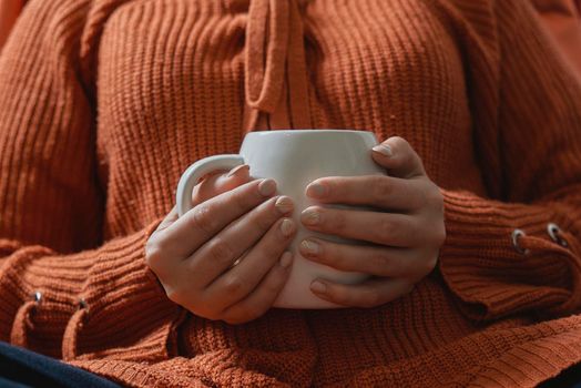 Autumn mood. Young female in orange woolen pullover drinking refreshing coffee in a cold autumnal morning. Woman in sweater holding in hands a cup of tea