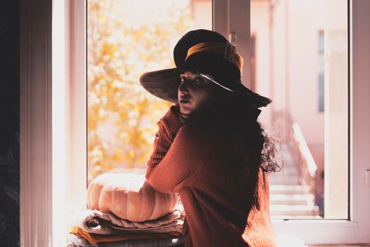 Halloween lady wearing witch cup. A woman in witch hat and Halloween pumpkin near the windows. Portrait of young female in orange sweater and traditional black witch cap