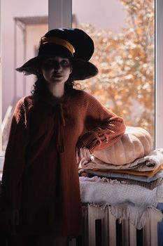 Halloween lady wearing witch cup. A woman in witch hat and Halloween pumpkin near the windows. Portrait of young female in orange sweater and traditional black witch cap