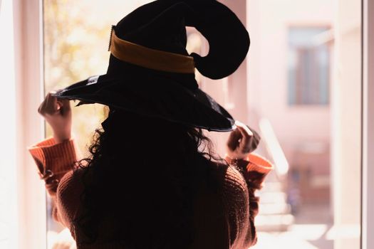 Halloween lady wearing witch cup. A woman in witch hat and Halloween pumpkin near the windows. Portrait of young female in orange sweater and traditional black witch cap