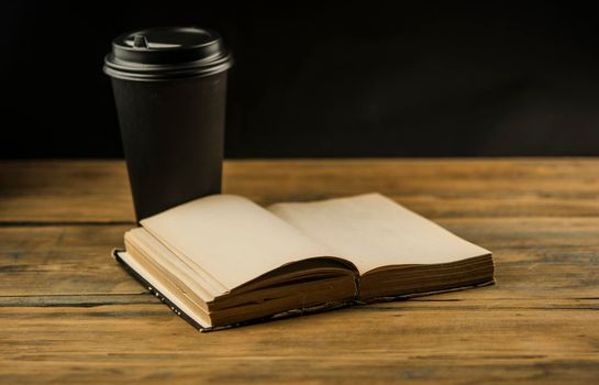 Open book and a cup of coffee in a disposable paper cup on a wooden table