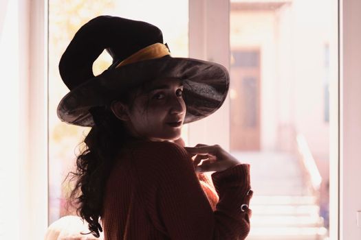 Halloween lady wearing witch cup. A woman in witch hat and Halloween pumpkin near the windows. Portrait of young female in orange sweater and traditional black witch cap