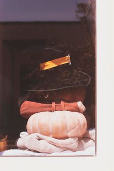 Halloween lady wearing witch cup. A woman in witch hat and Halloween pumpkin near the windows. Portrait of young female in orange sweater and traditional black witch cap