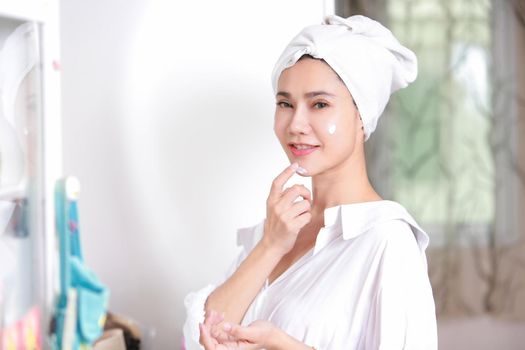 Young woman applying cream to face mirror reflection at dressing room.