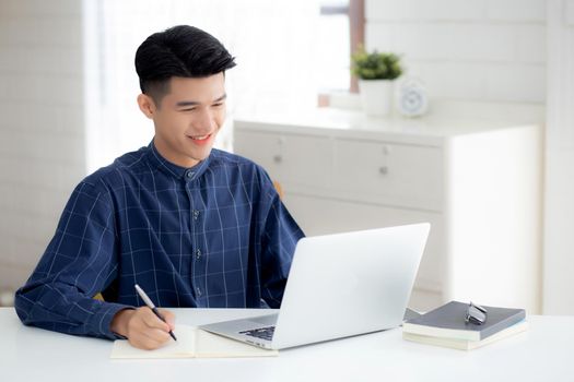 Young asian business man writing on notebook for planning working and using laptop computer on desk at home, notes about finance, male study and learning, business and communication concept.