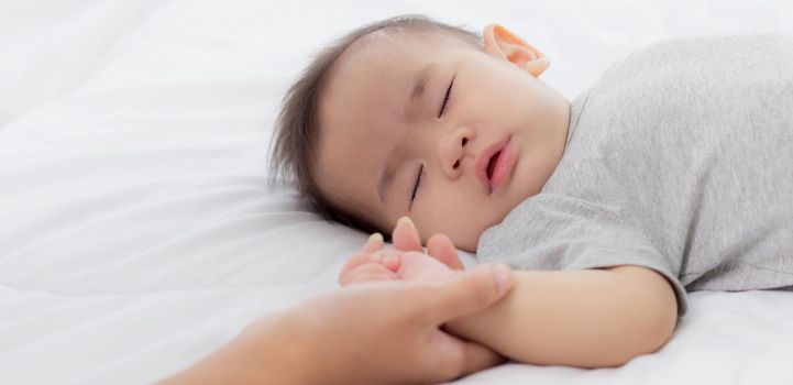 Young asian mother touch hand of little baby girl with tender on bed in the bedroom, mom love newborn and care, woman with expression with child together, parent and daughter, family concept.