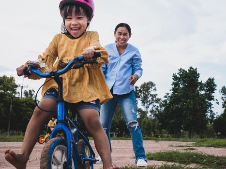 Cute little girl in safety helmet learn to ride a bike with her mother in summer park. Outdoor sports for kids. childhood happiness. family spending time together.