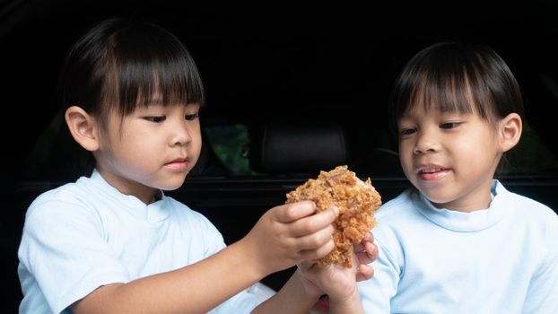 Two sibling girls having break during family road trip and eating fried chicken in trunk of car on the park. Delivery service and Social distance from Covid-19 or Coronavirus.