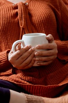 Autumn mood. Young female in orange woolen pullover drinking refreshing coffee in a cold autumnal morning. Woman in sweater holding in hands a cup of tea