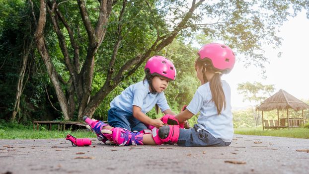 Cute older sister is helping younger sister to put on a protection pads and safety helmet practicing to roller skate on the street in the park. Active outdoor sport for kids.