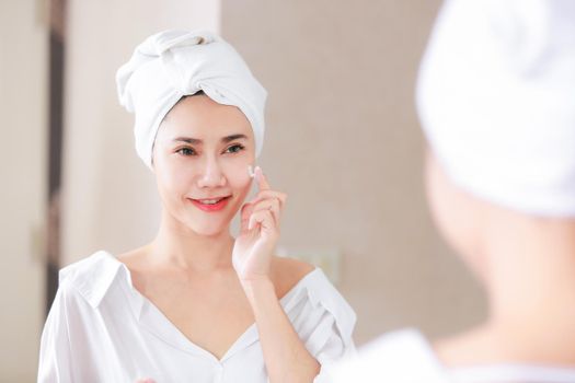 Young asia woman applying foundation or moisturizer on her face in front of mirror.