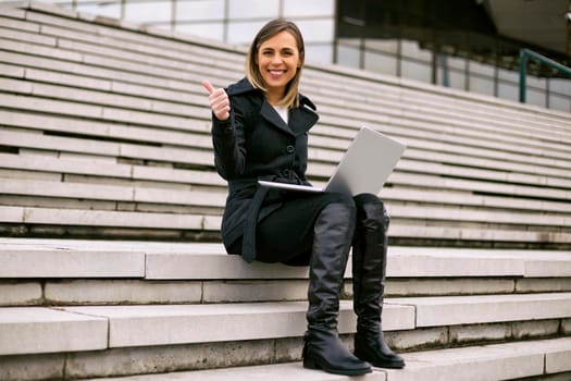 Beautiful businesswoman using laptop and showing thumb up while sitting on the staircase in the city.