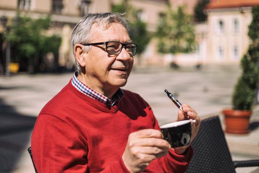 Cheerful senior man enjoys drinking coffee and smoking electronic cigarette at the bar.Image is intentionally toned.