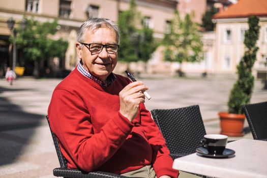 Cheerful senior man enjoys drinking coffee and smoking electronic cigarette at the bar.Image is intentionally toned.