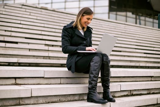 Beautiful businesswoman sitting on the staircase in the city and using laptop.
