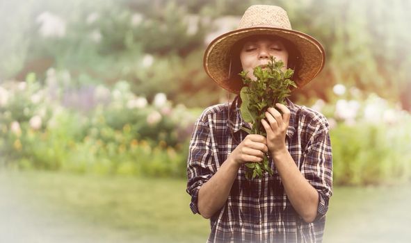 A young girl gardener in a straw hat holds a bouquet of harvested fresh mint and inhales its wonderful menthol scent, a woman is harvesting in the garden.