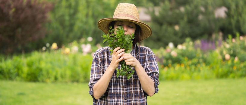 A young girl gardener in a straw hat holds a bouquet of harvested fresh mint and inhales its wonderful menthol scent, a woman is harvesting in the garden.