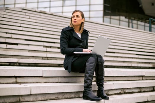 Beautiful businesswoman sitting on the staircase in the city and using laptop.