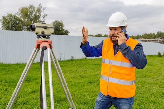 A disgruntled civil engineer with an optical level gauge speaks on a cell phone and swears. A builder in a helmet with a beard is talking on the phone with colleagues.
