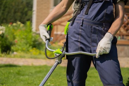 A close up of a young man moving a lawn with a lawn mower in his hands. A man is caring for his backyard garden. A professional gardener is cutting the grass, hands in gloves.