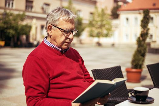 Senior man enjoys reading book and drinking coffee at the bar.