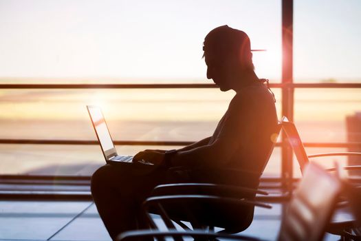 A young man is working on a laptop at the airport while waiting to board the plane. A man is engaged in business, buys tickets, studies and communicates via the Internet at sunset.