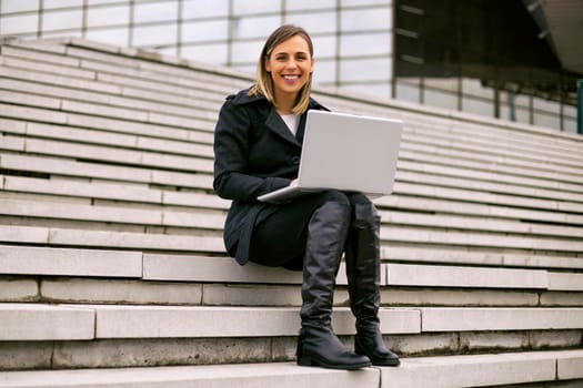 Beautiful businesswoman sitting on the staircase in the city and using laptop.