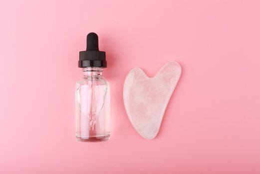 Flat lay with massage gel, skin serum or oil in transparent glass bottle and heart shaped guasha pink quartz crystal on bright pink background. Concept of facial massage and self treatment
