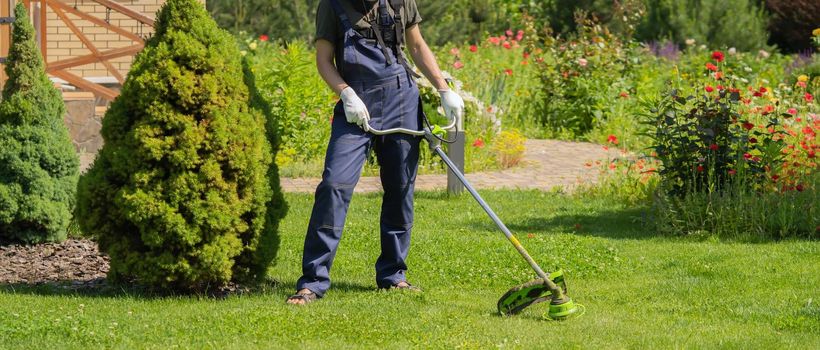A young man is mowing a lawn with a lawn mower in his beautiful green floral summer garden. A man with a lawnmower cares for the grass in the backyard.