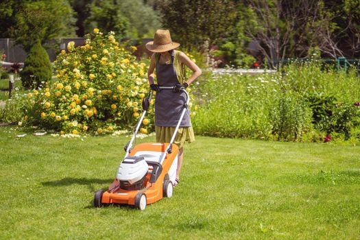 A young girl in a straw hat is mowing a lawn in the backyard with an orange lawn mower. A woman gardener is trimming grass with the grass cutter. A lawnmower is cutting a lawn on a summer sunny day.