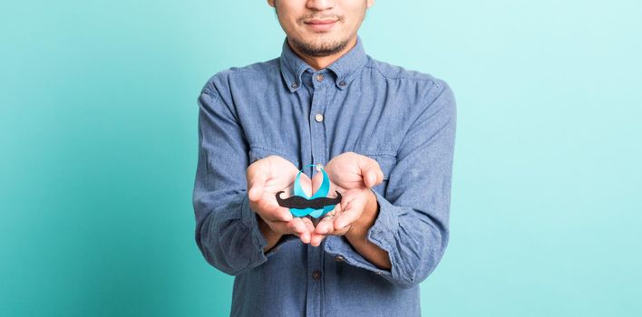 Asian portrait happy handsome man posing he holding a light blue ribbon and mustache, studio shot isolated on blue background, Prostate Cancer Awareness in November concept