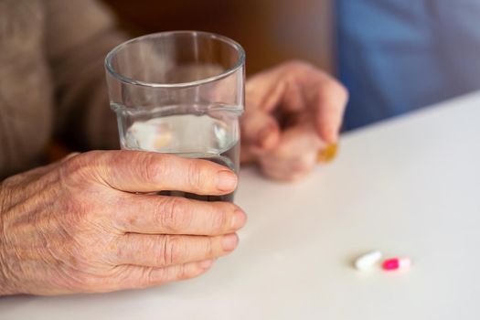An elderly woman holds a glass of water in her wrinkled hand to take a pill or vitamins. Treatment in old age, combating headaches, migraines, colds. Medicine and self-care.