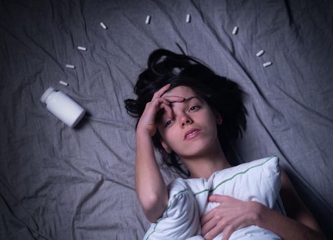 A young tired girl woke up in the middle of the night with a headache and insomnia, hugging a pillow and reaching out for sleeping pills. A woman with health problems needs medical attention.