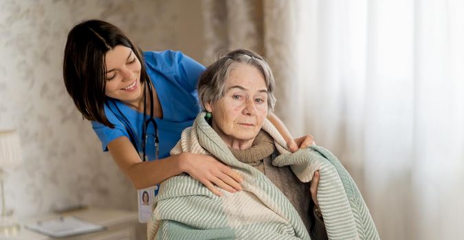 A young nurse takes care of an elderly 80-year-old woman at home, wraps a blanket around her. Happy retired woman and trust between doctor and patient. Medicine and healthcare.