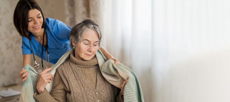 A young nurse takes care of an elderly 80-year-old woman at home, wraps a blanket around her. Happy retired woman and trust between doctor and patient. Medicine and healthcare.