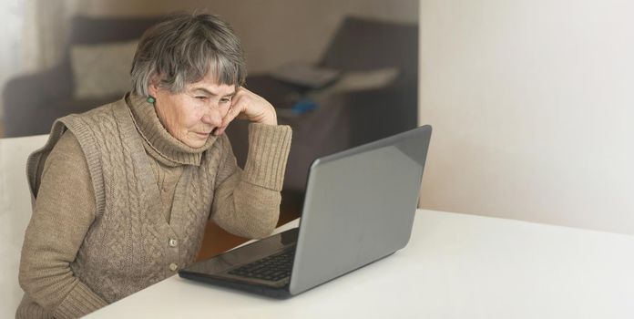 Old woman wearing casual clothes such as knitted sweater, spending free time at cozy home, sitting, resting using pc, typing message and searching, reading news online.