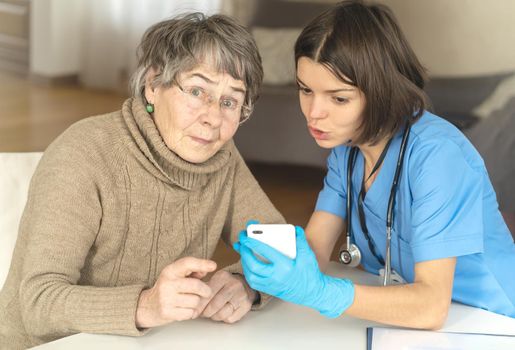 A nurse in a medical suit takes care and explains to an elderly patient how to use applications on a smartphone. Grandmother 80 years old, does not understand how to communicate with a doctor online.