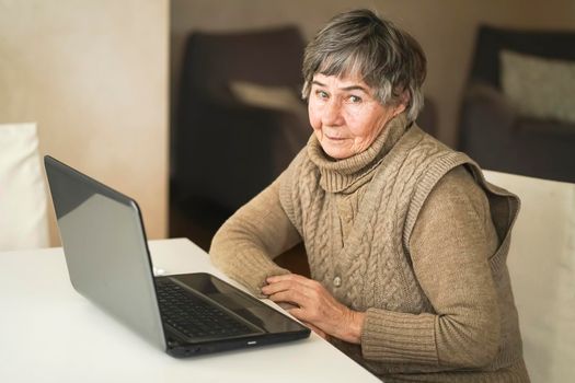 Old smiley woman wearing casual clothes such as knitted sweater, spending free time at cozy home, sitting, resting using pc, typing message and searching, reading news online.
