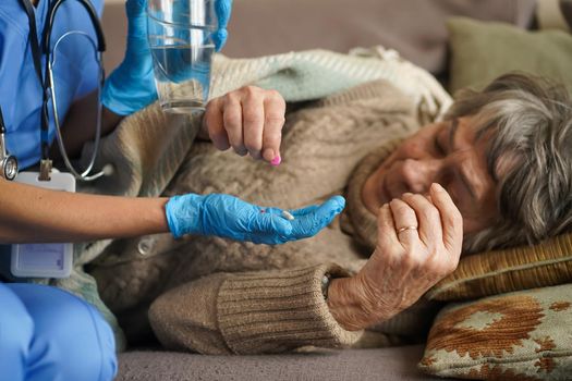 A young nurse is caring for an elderly 80-year-old woman at home. She holds a glass of water and gives medicine pills a pensioner retired woman who lies and rests in bed.