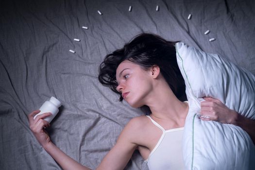A young tired girl woke up in the middle of the night with a headache and insomnia, hugging a pillow and reaching out for sleeping pills. A woman with health problems needs medical attention.