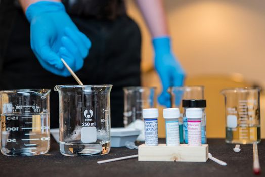 A chemistry experiment with a young scientist wearing latex gloves using liquid filled beakers and various vials and stirring mechanisms.