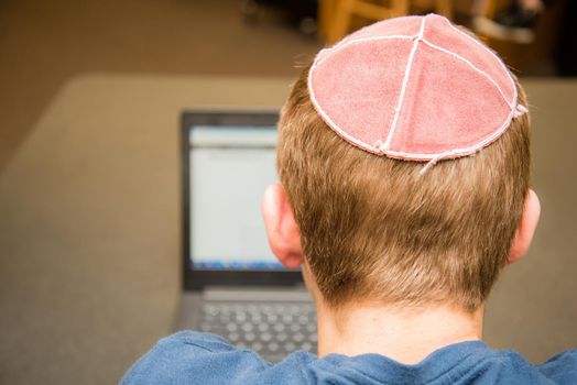Young man wearing a yarmulke from the back doing work on a laptop in a library with colorful books on shelves.