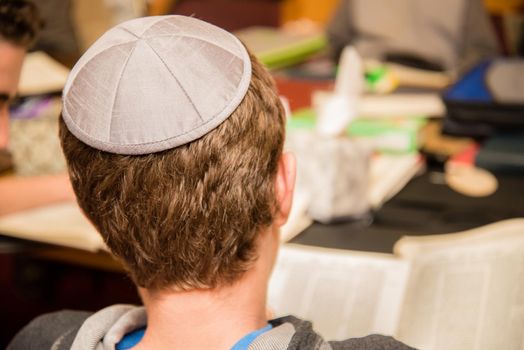 Young Jewish boy wearing yarmulke from the back sitting in a classroom setting with students.